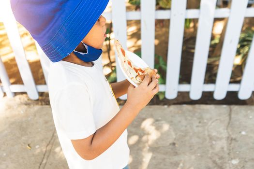 Latin boy enjoying the summer and eating pizza dressed in comfortable clothes and a blue bucket hat