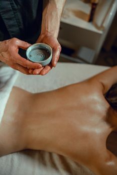 A bowl with warm herb infused oil for back massage in hands of therapist. Woman getting massage