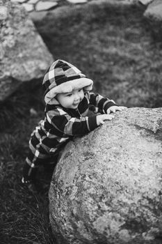 Portrait of cute baby in checked green, blue and black overall with hood sitting on the ground covered with bright dry foliage on autumn day. Fair haired baby in overall on vivid colorful leaves in the autumnal park or forest.