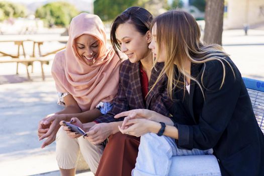 Content young multiracial female best friends, in casual clothes and hijab, smiling while watching video on smartphone sitting on bench in city park on sunny day