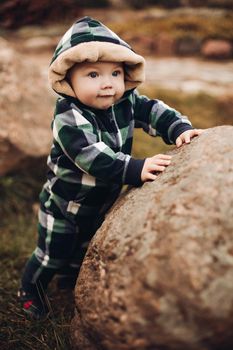 Portrait of cute baby in checked green, blue and black overall with hood sitting on the ground covered with bright dry foliage on autumn day. Fair haired baby in overall on vivid colorful leaves in the autumnal park or forest.