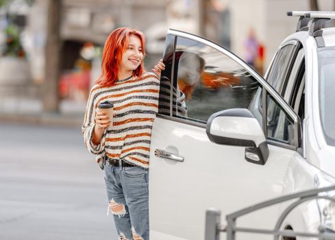 Pretty girl with coffee cup standing close to car at street and smiling. Stylish trendy teenager with bright color of hair portrait outdoors
