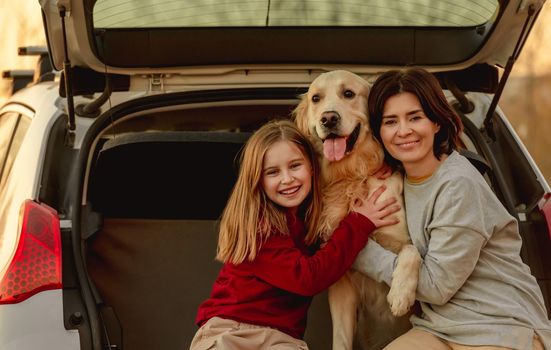 Family girl and child hugging golden retriever dog sitting in car trunk outdoors. Young mother and daughter kid with doggy pet labrador in vehicle