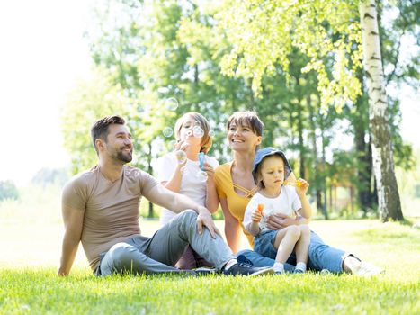 Happy family of mother father and two children playing with soap bubble on meadow