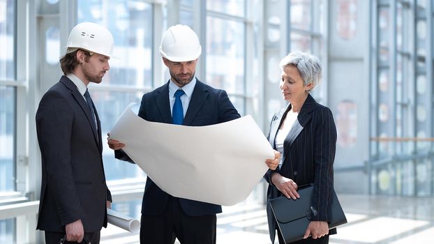 Multi ethnic business architect people in safety helmets discussing blueprint standing in the lobby at office