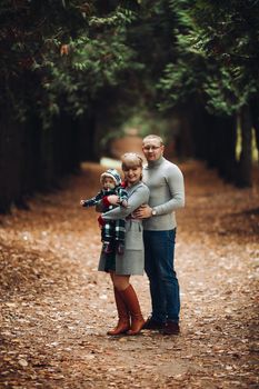 Portrait of attractive young mother and handsome smiling father wearing glasses holding their beautiful lovely baby girl on hands standing against green hedge in autumnal park. They are smiling and looking at camera.