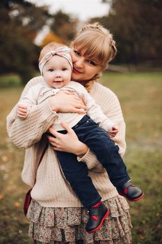 Professional portrait of attractive young woman with fair hair in braid embracing her little baby girl in plaid warm overall while standing in beautiful bright tree with red leaves. They are smiling at camera surrounded by vivid foliage in autumnal park.