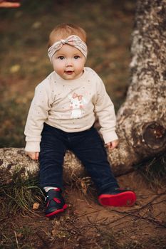 Portrait of lovely little baby girl wearing cute jersey with bunny on it and beige patterned bow headband. She is smiling at camera while parents holding her arms. Weekend in the park in autumn. Baby doing her first steps.