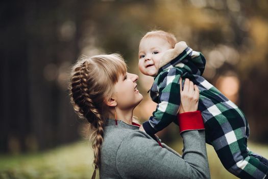 Professional portrait of attractive young woman with fair hair in braid embracing her little baby girl in plaid warm overall while standing in beautiful bright tree with red leaves. They are smiling at camera surrounded by vivid foliage in autumnal park.