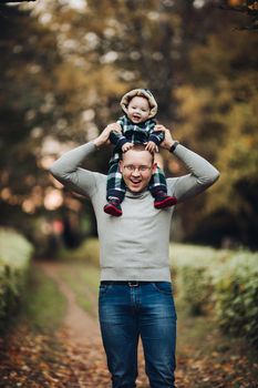 Professional portrait of attractive young man embracing his little baby girl in plaid warm overall while standing in beautiful bright tree with red leaves. They are smiling at camera surrounded by vivid foliage in autumnal park.