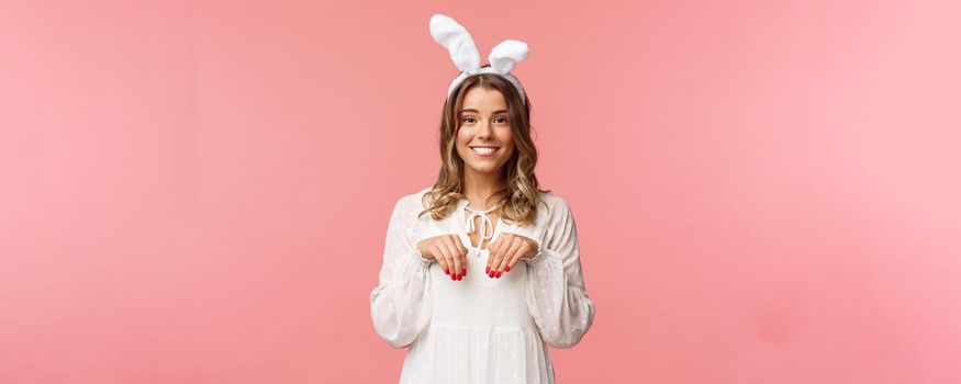 Holidays, spring and party concept. Portrait of cute and tender, lovely blond girl imitating bunny, holding hands like paws and wearing rabbit ears, smiling camera, pink background.