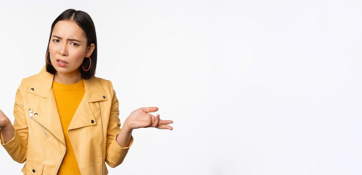 Image of confused young asian woman shrugging shoulders, looking puzzled and clueless at camera, cant understand, standing against white background.