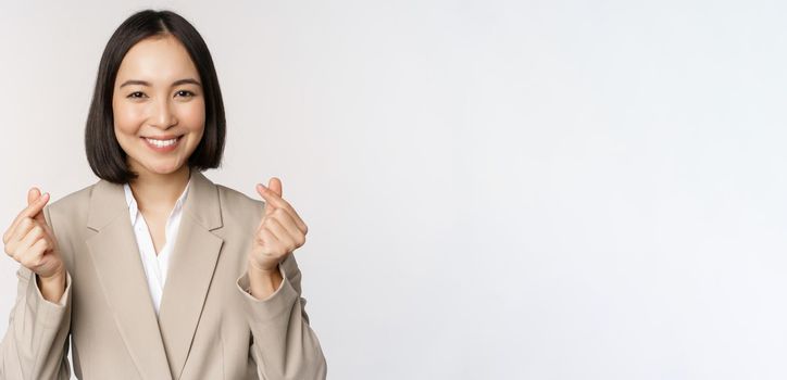 Cheerful asian saleswoman, smiling and showing finger hearts sign, standing in suit over white background.