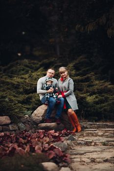 Portrait of attractive young mother and handsome smiling father wearing glasses holding their beautiful lovely baby girl on hands standing against green hedge in autumnal park. They are smiling and looking at camera.