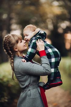 Professional portrait of attractive young woman with fair hair in braid embracing her little baby girl in plaid warm overall while standing in beautiful bright tree with red leaves. They are smiling at camera surrounded by vivid foliage in autumnal park.