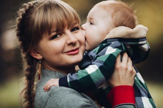 Professional portrait of attractive young woman with fair hair in braid embracing her little baby girl in plaid warm overall while standing in beautiful bright tree with red leaves. They are smiling at camera surrounded by vivid foliage in autumnal park.