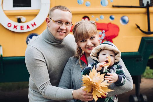 Portrait of attractive young mother and handsome smiling father wearing glasses holding their beautiful lovely baby girl on hands standing against green hedge in autumnal park. They are smiling and looking at camera.