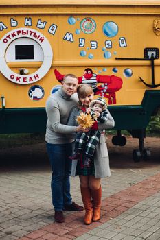 Portrait of attractive young mother and handsome smiling father wearing glasses holding their beautiful lovely baby girl on hands standing against green hedge in autumnal park. They are smiling and looking at camera.