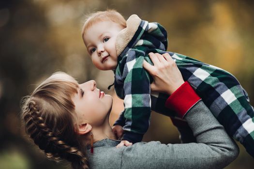 Professional portrait of attractive young woman with fair hair in braid embracing her little baby girl in plaid warm overall while standing in beautiful bright tree with red leaves. They are smiling at camera surrounded by vivid foliage in autumnal park.