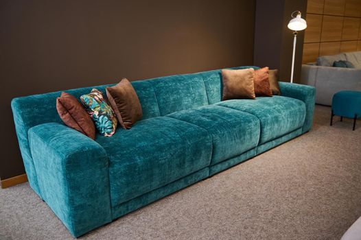 Exhibition of modern stylish upholstered furniture in the showroom of a furniture store. Focus on a turquoise soft velour sofa and brown cushions lit by a lamp against a brown wall background