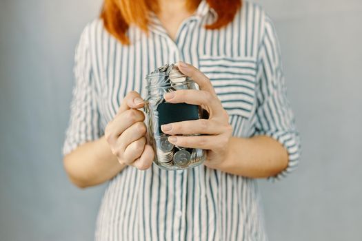 Glass bottle with silver coins in women's hands. Blank black sticker on donation money jar. Redhead girl n white and black striped shirt holds her savings. Pot for tips.