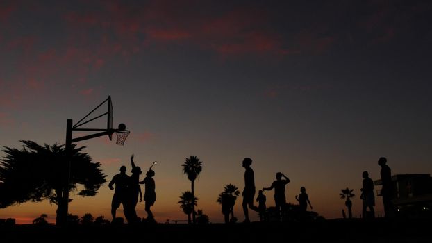 People playing basket ball game, silhouettes of players on basketball court outdoor, sunset ocean beach, California coast, Mission beach, USA. Black hoop, net and backboard on streetball sport field.