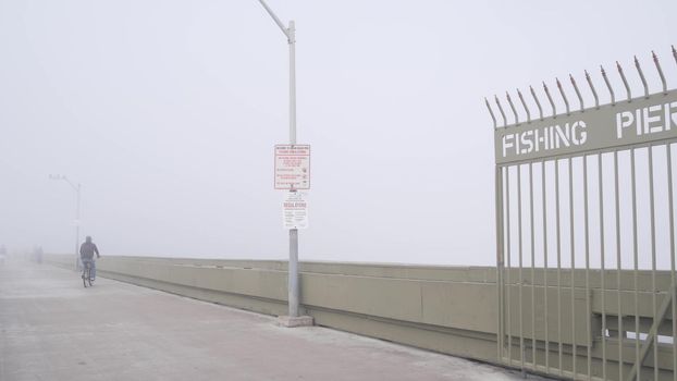 Metal gate on Ocean Beach pier in fog, misty California coast, USA. Foggy moody cloudy weather on San Diego shore. Calm tranquil atmosphere. Waterfront boardwalk entrance in gloomy depressive haze.