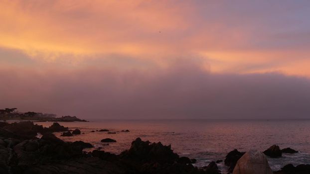 Rocky craggy ocean beach, calm sea waves, Monterey bay, 17-mile drive seascape, California coast, USA. Beachfront waterfront Pacific Grove, waterside promenade. Pink purple pastel dramatic sunset sky.