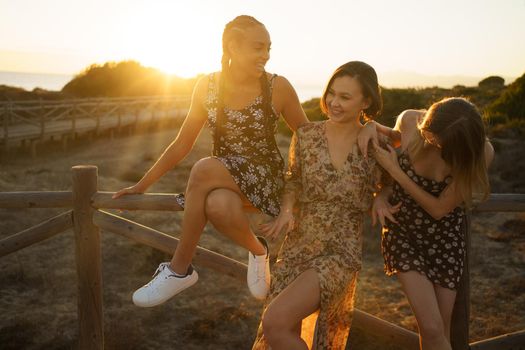 Delighted multiracial female friends in sundresses standing near wooden barrier on waterfront on sunny summer day in coastal area against cloudless sky