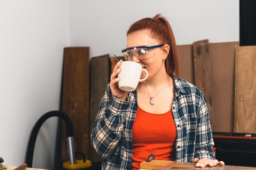 Young red-haired woman carpenter, concentrated and smiling, working on the layout of wood in a small carpentry shop, dressed in a blue checked shirt and a red t-shirt. Woman carpenter having a coffee, in the morning in her small carpentry business. Warm light indoors, background with wooden slats. Horizontal.