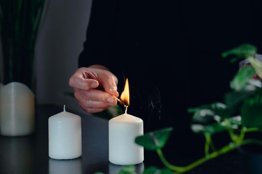 Close-up of the hands of a mature beautician, dressed in black company uniform, lighting white scented candles in her small spa business, to start the day. dim indoor lighting. Horizontal