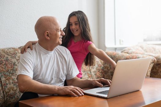 Happy little girl hugging smiling grandfather sitting on sofa with laptop