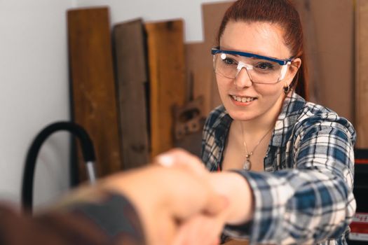 Young red-haired woman carpenter, concentrated and smiling, working on the layout of wood in a small carpentry shop, dressed in a blue checked shirt and a red t-shirt. Woman carpenter talking to a customer and shaking hands to close a deal. Warm light indoors, background with wooden slats. Horizontal.