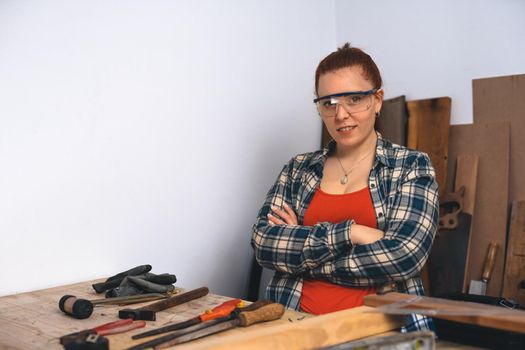 Close-up of the hands of a young red-haired carpenter woman, concentrating and smiling, working in her small carpentry workshop, dressed in a blue checked shirt and a red t-shirt. Woman carpenter using posing for camera in her carpentry shop. Warm light indoors, background with wooden slats. Horizontal. Looking at the camera.