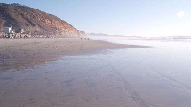 Steep cliff, rock or bluff, California coast erosion, Del Mar, San Diego, USA. People walking along eroded crag on littoral sand. Torrey Pines state ocean beach, low tide water waves. Foggy weather.