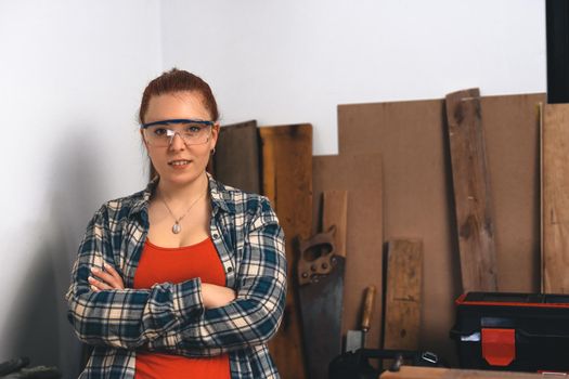 Close-up of the hands of a young red-haired carpenter woman, concentrating and smiling, working in her small carpentry workshop, dressed in a blue checked shirt and a red t-shirt. Woman carpenter using posing for camera in her carpentry shop. Warm light indoors, background with wooden slats. Horizontal. Looking at the camera.