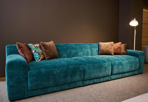 Exhibition of modern stylish upholstered furniture in the showroom of a furniture store. Focus on a turquoise soft velour sofa and brown pillows lit by a lamp against a brown wall background