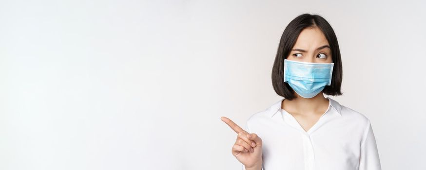 Image of young asian woman in medical face mask, pointing left and looking with suspicious confused expression, standing over white background.