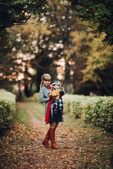 Professional portrait of attractive young woman with fair hair in braid embracing her little baby girl in plaid warm overall while standing in beautiful bright tree with red leaves. They are smiling at camera surrounded by vivid foliage in autumnal park.