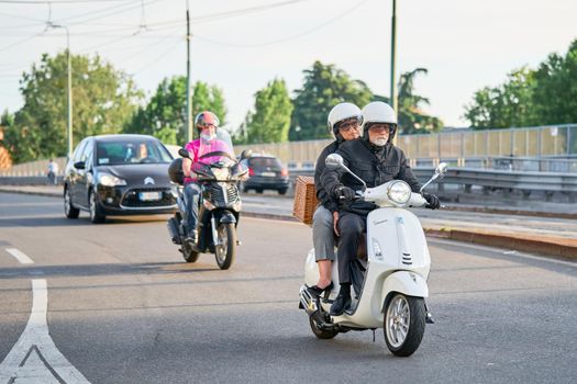 Senior couple riding motorbike in busy city street traffic. Motorcyclists driving motorcycle and cars on the road. Commuting. Milan, Italy - May 26, 2020