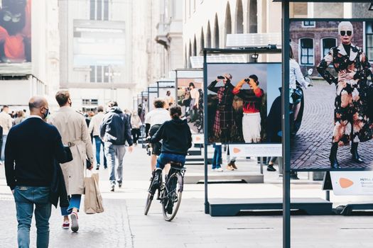 Milan, Italy - September 27, 2020. People in masks walking street in downtown of Milan, commuting, cycling. New normal during covid. Modern fashion photography street exhibition using solar batteries