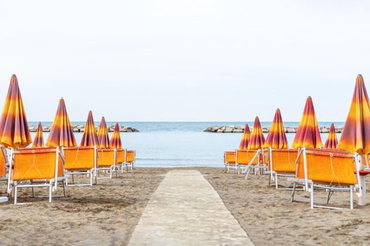 Lounge chairs on sand beach in Italy. Beach lounge area with orange umbrellas and green sunbeds. View on ocean and horizon. Sunbeds and parasols on the seashore. Adriatic coast, Rimini