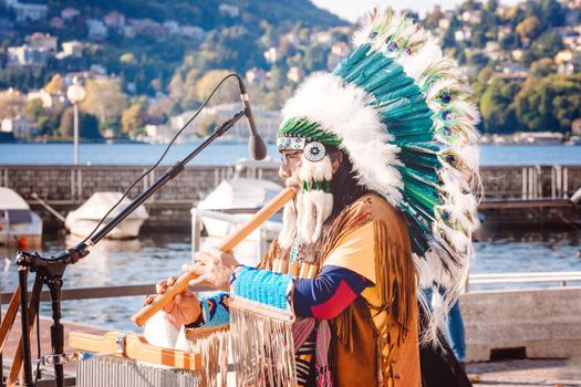 American Indian musician performing on the street in traditional costume in feather headdress, Como lake, Italy. Artist plays music and sings in the street for tourists. Como, Italy - 10 October, 2019