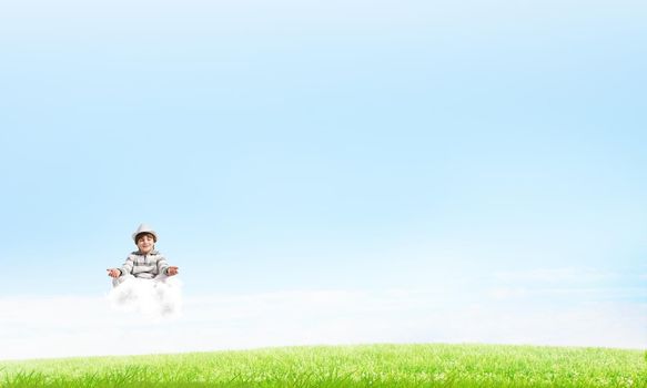 Young little boy keeping eyes closed and looking concentrated while meditating on cloud in the air with bright and beautiful landscape on background.