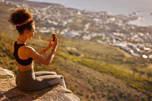 Shot of a woman taking pictures with her cellphone while sitting on a mountain cliff.