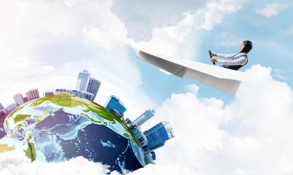 Businessman in aviator hat sitting in paper plane and holding steering wheel. Pilot driving paper plane in cloudy blue sky. Spherical view of modern city with high skyscrapers and sea line.