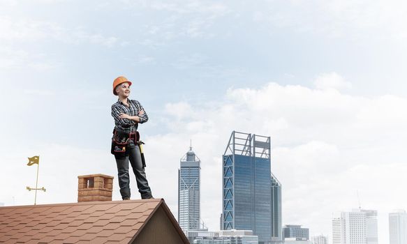 Attractive female construction worker in hardhat standing with folded arms. Confident young woman in checkered blue shirt standing on brick roof of building. Professional perspective and motivation