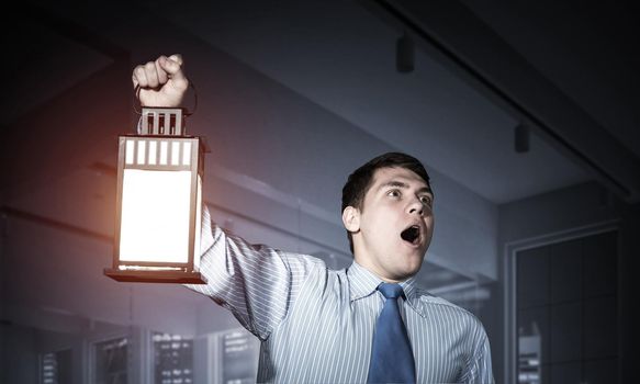 Horrified businessman holding glowing lantern on background office interior. Emotional corporate employee in shirt and tie looking for something at night. Shocked man with open mouth lost in dark