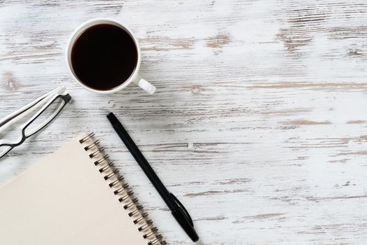 Top view office desk with white cup of coffee. Flat lay vintage wooden table with spiral notebook, pen and glasses. Coffee break time at work. Corporate business concept with copy space.