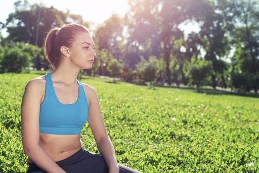 Beautiful smiling girl in activewear relax in park at sunrise. Portrait of young charming woman sitting on green grass. Training and meditation outdoor. Morning exercises and healthy lifestyle.